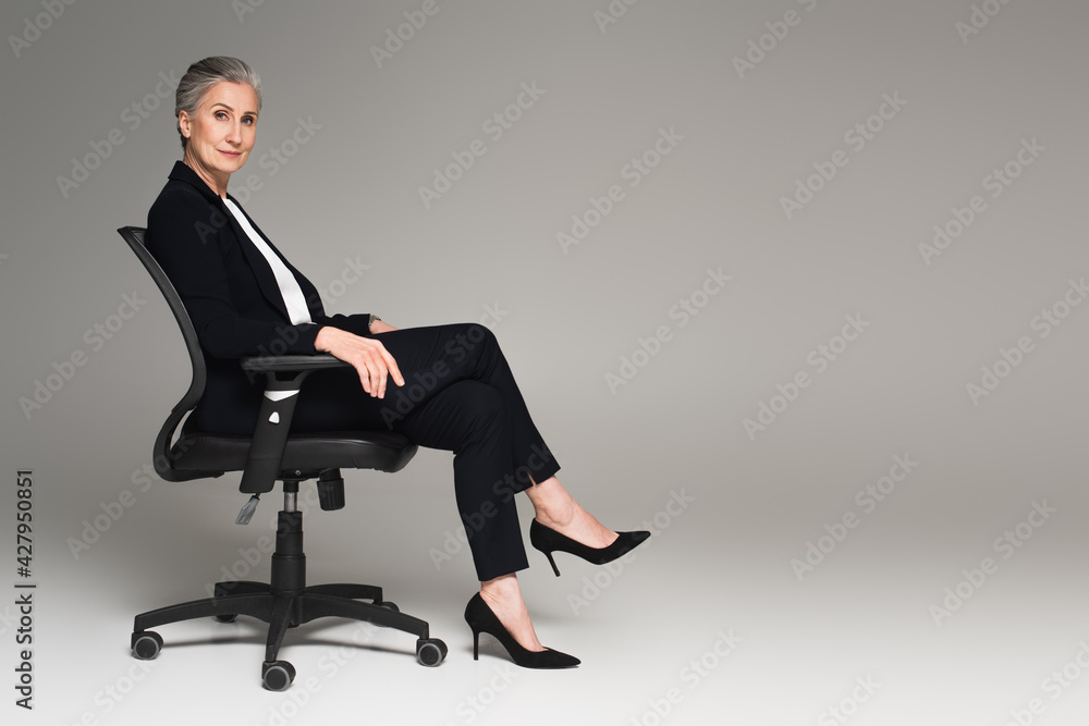 Middle aged businesswoman with crossed legs looking at camera on office chair on grey background Middle aged businesswoman with crossed legs looking at camera on office chair on grey background