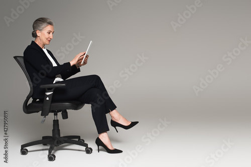 Foto Mature businesswoman using digital tablet on office chair on grey background wit