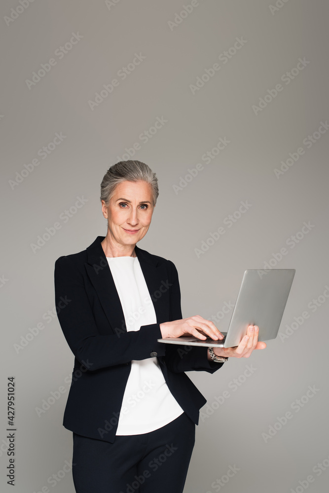 Mature businesswoman using laptop and looking at camera isolated on grey Mature businesswoman using laptop and looking at camera isolated on grey