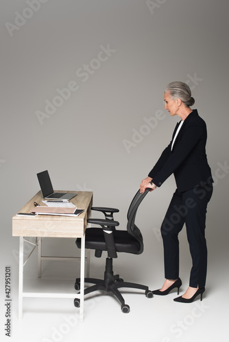 Side view of mature businesswoman standing near office chair and working table on grey background
