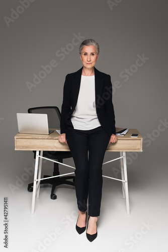 Mature businesswoman looking at camera near gadgets on table on grey background