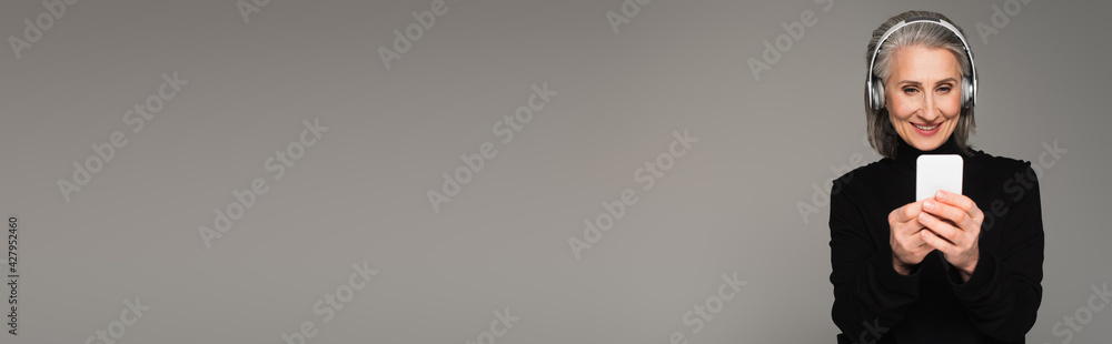 Smiling woman in black turtleneck using cellphone and headphones isolated on grey, banner Smiling woman in black turtleneck using cellphone and headphones isolated on grey, banner