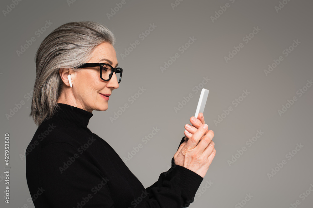 Mature woman in earphone using smartphone isolated on grey Mature woman in earphone using smartphone isolated on grey