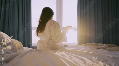 Tranquil young woman in white bathrobe drinks coffee sitting on large comfortable bed near panoramic window in hotel room at sunrise backside view