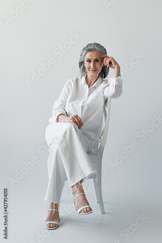 Smiling mature woman sitting with crossed legs on chair on grey background