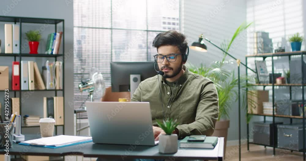 Young handsome Hindu man in glasses and headphones with microphone sitting in office alone and working on laptop. Young handsome Hindu man in glasses and headphones with microphone sitting in office alone and working on laptop.