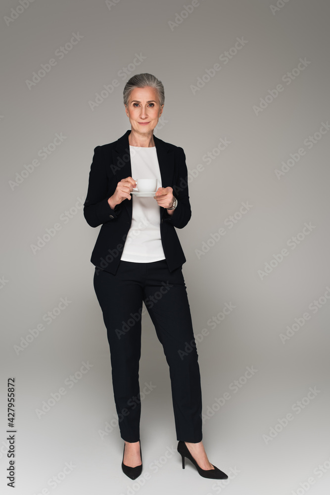 Senior businesswoman with cup and saucer looking at camera on grey background Senior businesswoman with cup and saucer looking at camera on grey background