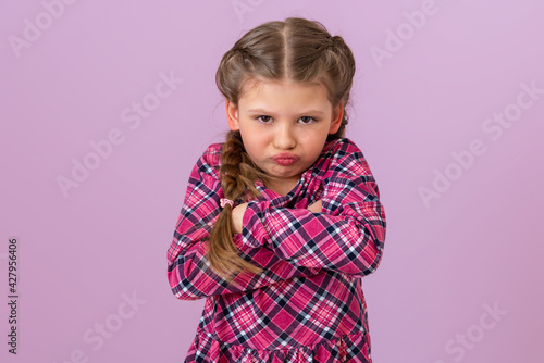 A little girl in a plaid dress and with pigtails on her head holds her hands near her chest and looks very offended.