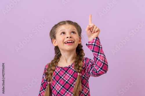 A little girl in a purple checkered dress points her finger up.