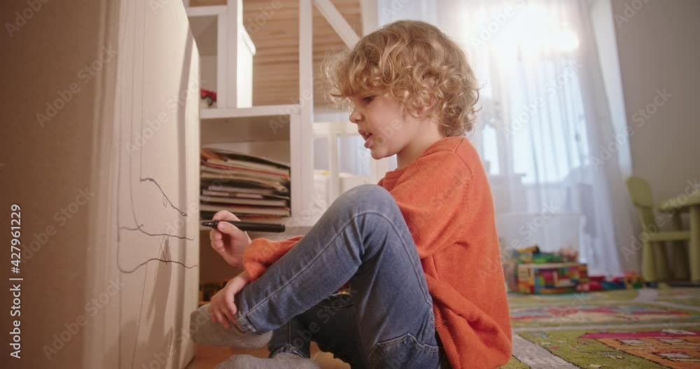 A preschool boy with curly hair drawings with a marker on a cardboard box dreams and is creative, sitting on the ground in a cozy children's room A preschool boy with curly hair drawings with a marker on a cardboard box dreams and is creative, sitting on the ground in a cozy children's room