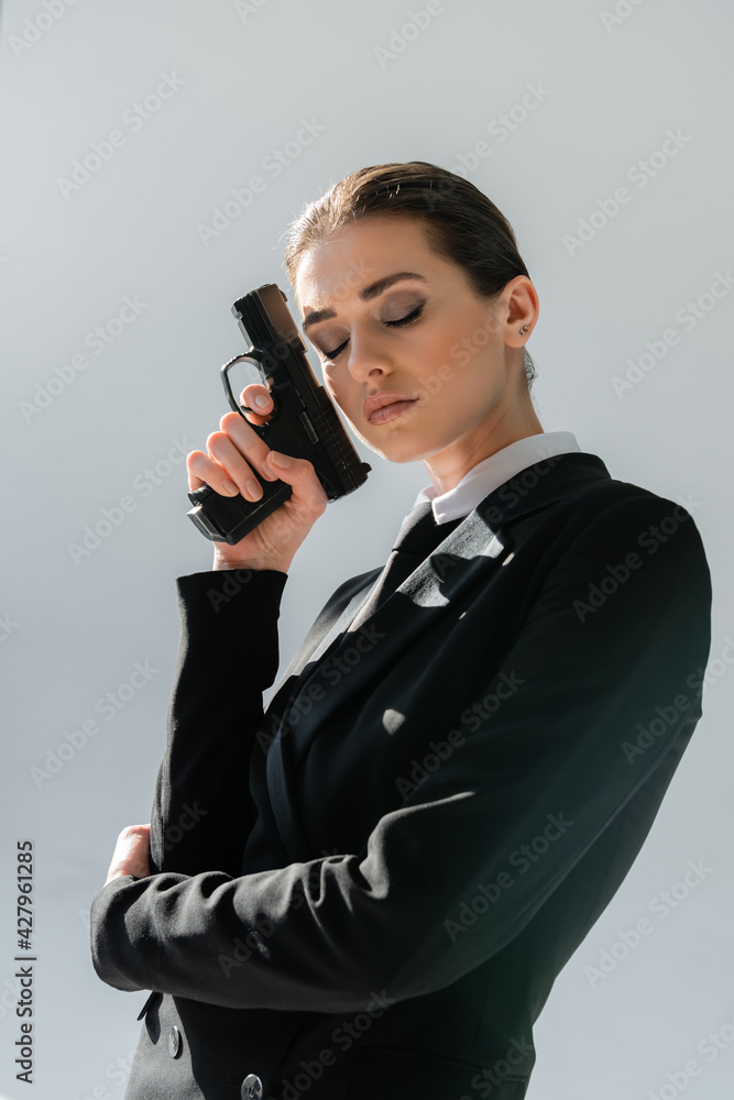 elegant woman holding gun near face while standing with closed eyes isolated on grey elegant woman holding gun near face while standing with closed eyes isolated on grey
