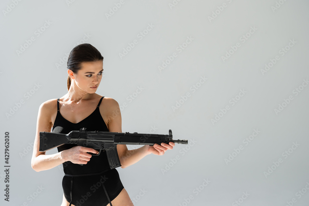 sexy woman in black bodysuit aiming with rifle isolated on grey