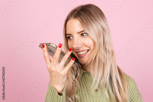 Pretty european woman in casual sweater on pink background holding mobile phone recording audio voice message happy smile, having conversation, excited cheerful