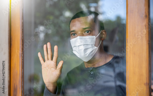Portrait of lonely African black man in medical mask looking through the window. Sad stressful man hand on glass isolated at home self quarantine. Prevent COVID-19. Coronavirus outbreak stay home