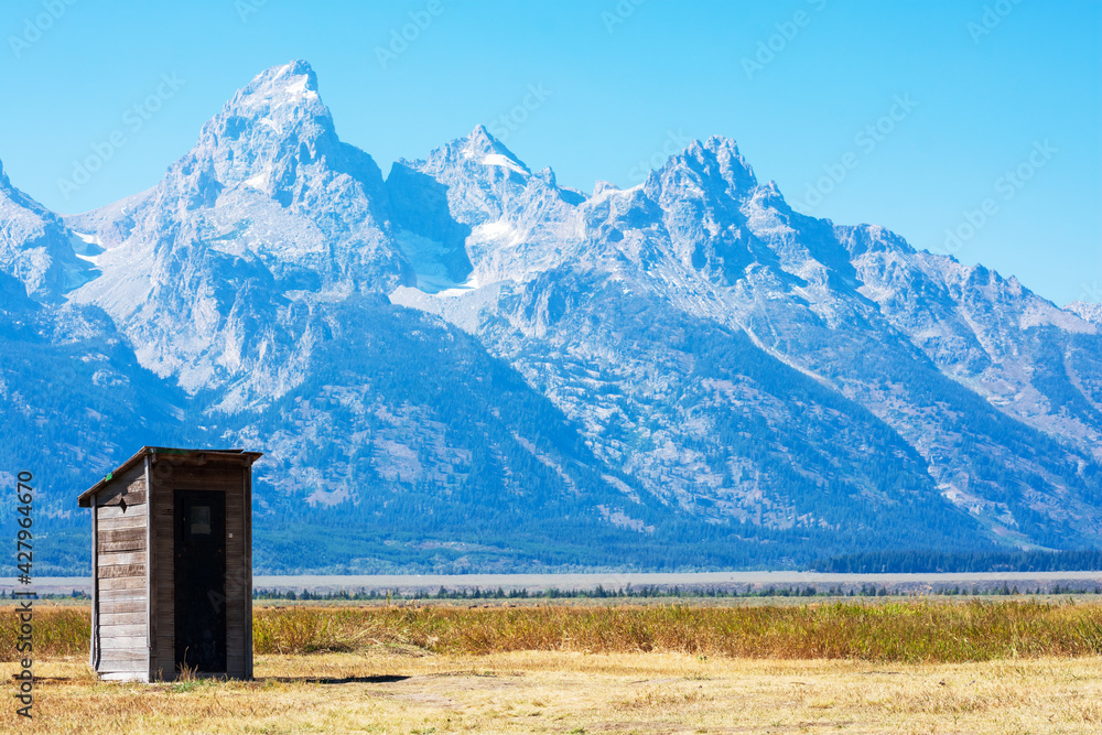 Old abandoned wooden outhouse at Mormon Row Historic District in scenic Antelope Flats valley area. Background majestic peaks of Teton Mountain Range in Grand Teton National Park.