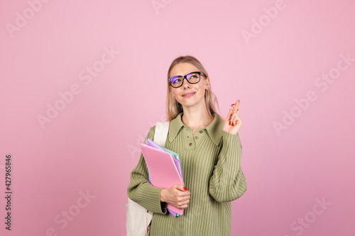 Pretty european woman in casual sweater on pink background with notebooks wait for special moment, keep fingers crossed, making wish gesture education concept