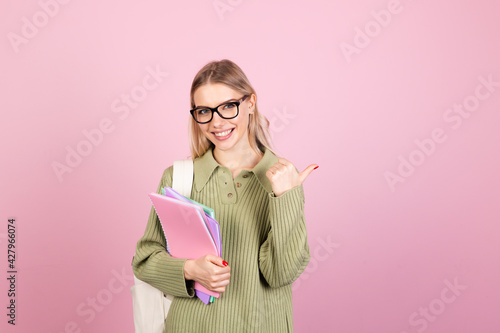 Pretty european woman in casual sweater on pink background with notebooks smiling happy pointing thumb finger right education concept
