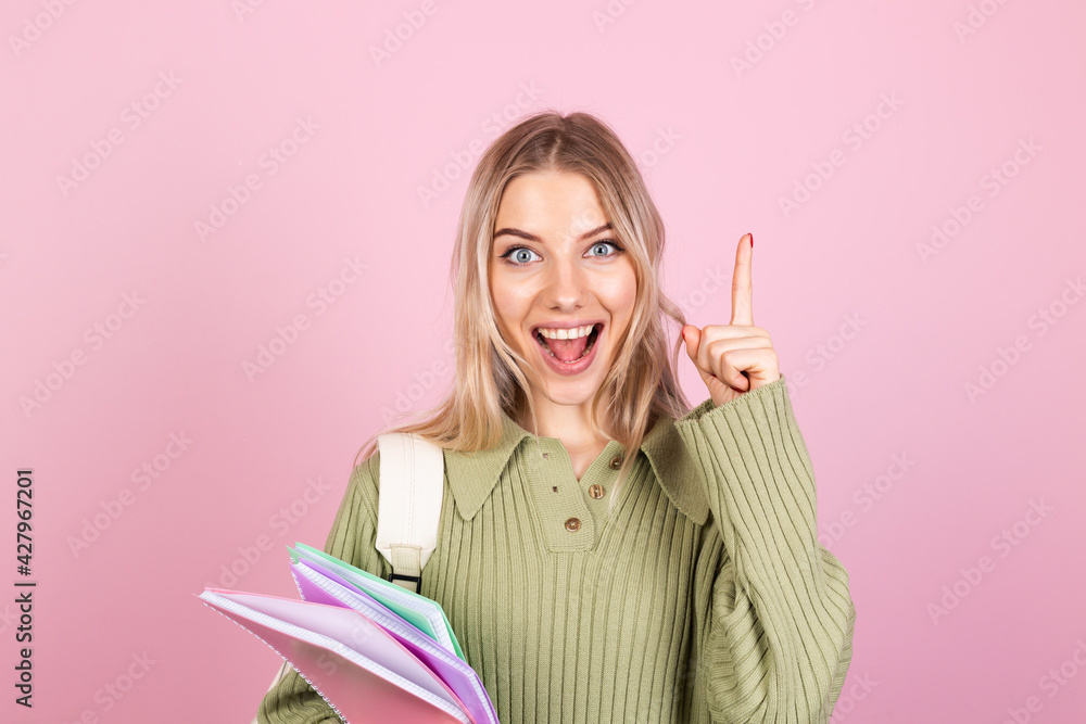 Pretty european woman in casual sweater on pink background with notebooks excited pointing finger up, idea came up, education high school concept