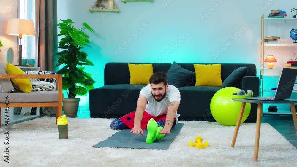 A handsome middle aged man with a dark brown beard is sitting on his workout mat in a room, he is continuo