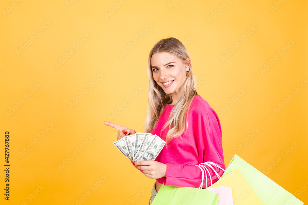 Pretty european woman in pink  blouse on yellow background smile excited hold fan of 100 dollars money and shopping bags, point index finger to left