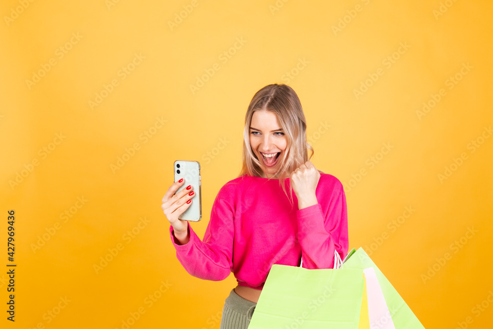 Pretty european woman in pink blouse on yellow background holding shopping bags doing winner gesture with mobile phone, discount sale concept Pretty european woman in pink blouse on yellow background holding shopping bags doing winner gesture with mobile phone, discount sale concept