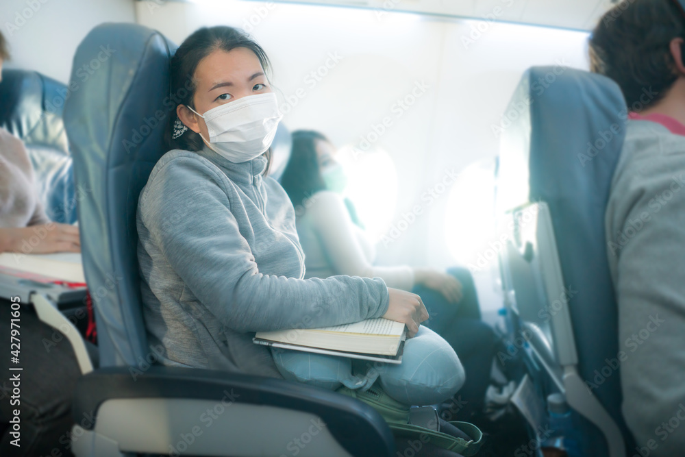flying in times of covid19 - young sweet and cute Asian Chinese woman in face mask sitting on airplane cabin reading book or novel enjoying the flight