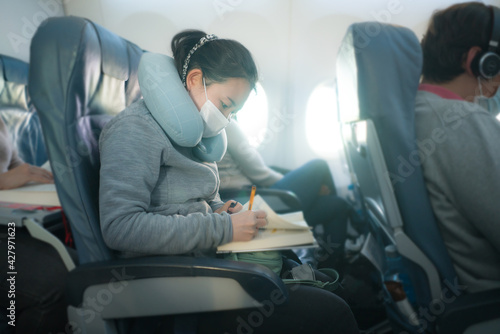 flying in times of covid19 - young sweet and cute Asian Korean woman in face mask sitting on airplane cabin reading book or novel enjoying the flight