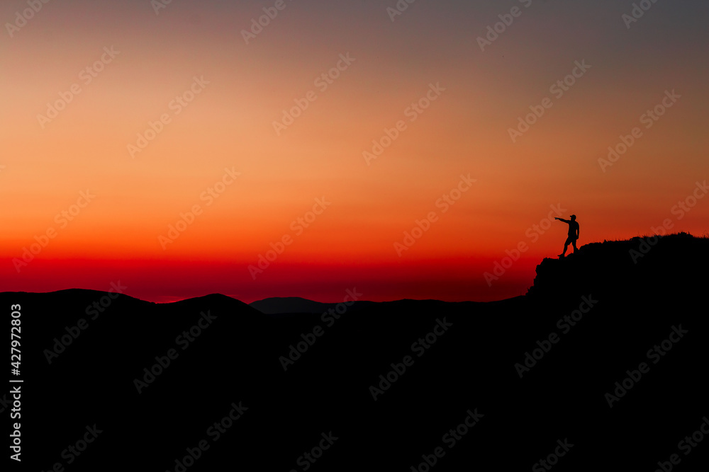 Red sunset with mountain silhouette. A man on mountain in sunset. Traveler on rock waving hands. Red sunset with mountain silhouette. A man on mountain in sunset. Traveler on rock waving hands.