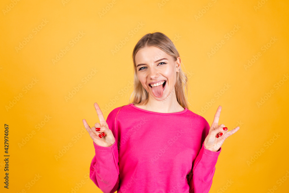 Pretty european woman in pink blouse on yellow background sticking tongue out doing rock symbol with hands up Pretty european woman in pink blouse on yellow background sticking tongue out doing rock symbol with hands up
