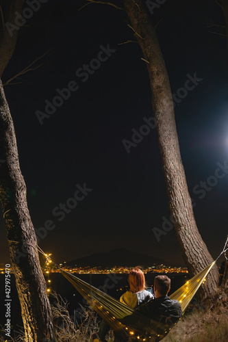 Couple relaxing in a hammock and enjoying starry sky. View from the top of the mountain to the lights of the city of Naples. Volcano Vesuvio. Sea and romance. Vertical photo. Sorrento Italy photo