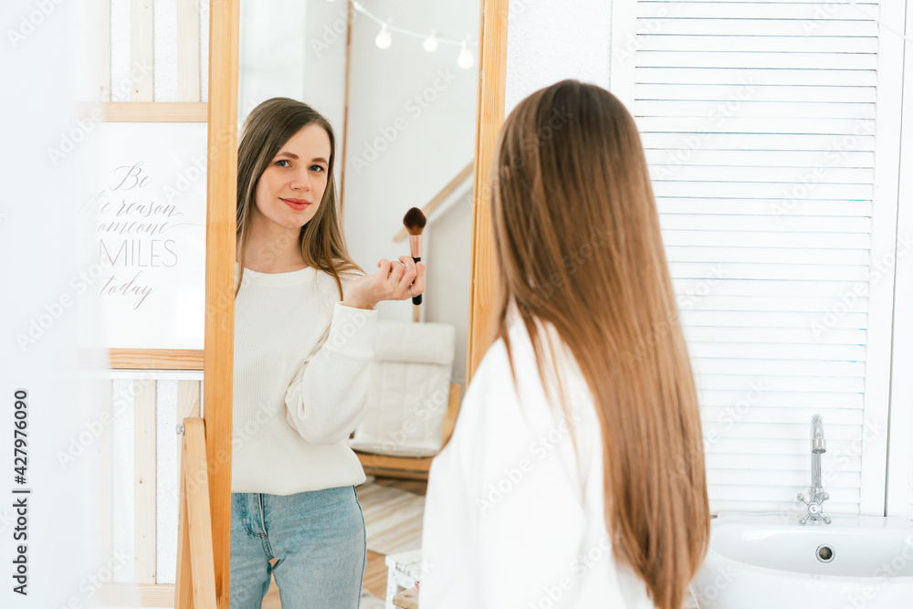 Beautiful smiling young caucasian woman with long hair standing near mirror, doing makeup and looking at reflection in living room at home. Beautiful smiling young caucasian woman with long hair standing near mirror, doing makeup and looking at reflection in living room at home.