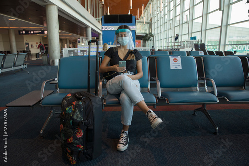 Covid-19 and social distancing. Traveling during pandemic. Young woman wearing a protective mask, waiting in the airport while looking at her smartphone. photo