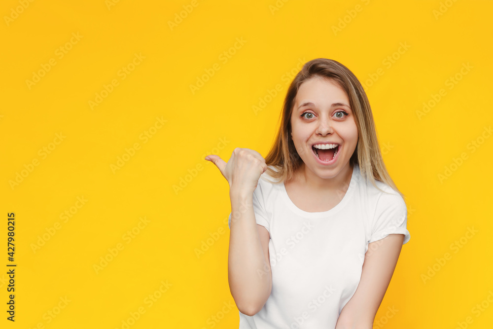 A young caucasian impressed smiling blonde woman in a white t-shirt indicate copy empty blank space for text or design with her finger presenting product isolated on a bright color yellow background A young caucasian impressed smiling blonde woman in a white t-shirt indicate copy empty blank space for text or design with her finger presenting product isolated on a bright color yellow background
