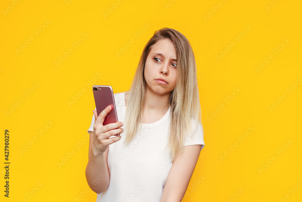 A frightened young caucasian blonde woman in a white t-shirt with a mobile phone in her hand looks suspiciously at the incoming call from an unknown number on a bright color yellow background. Fraud A frightened young caucasian blonde woman in a white t-shirt with a mobile phone in her hand looks suspiciously at the incoming call from an unknown number on a bright color yellow background. Fraud