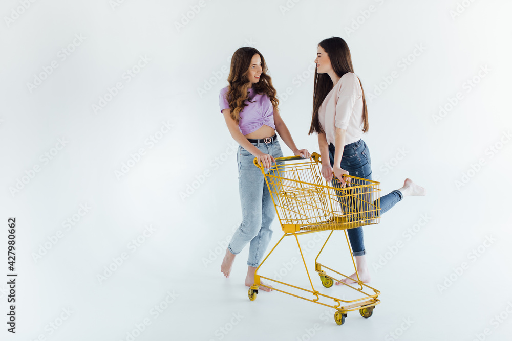 Side view of excited young friends smiling and raising hands while riding shopping trolley against white background