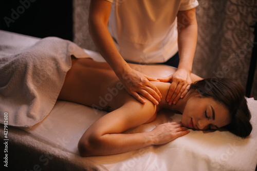 Middle shot portrait of young beauty woman lying on massage table with closed eyes. Professional massage therapist massages the shoulder of girl lying in spa centre, close-up. Concept of body care.