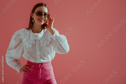 Photographie Fashionable  happy smiling girl wearing trendy green rectangular sunglasses, white vintage cotton blouse with lace collar, fuchsia color jeans, posing on pink background