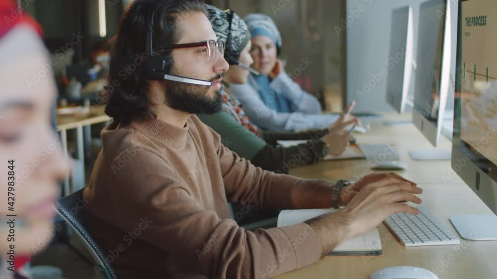 Side view selective focus shot of young middle eastern man in wireless headset talking on online call and using computer while working in support center Side view selective focus shot of young middle eastern man in wireless headset talking on online call and using computer while working in support center