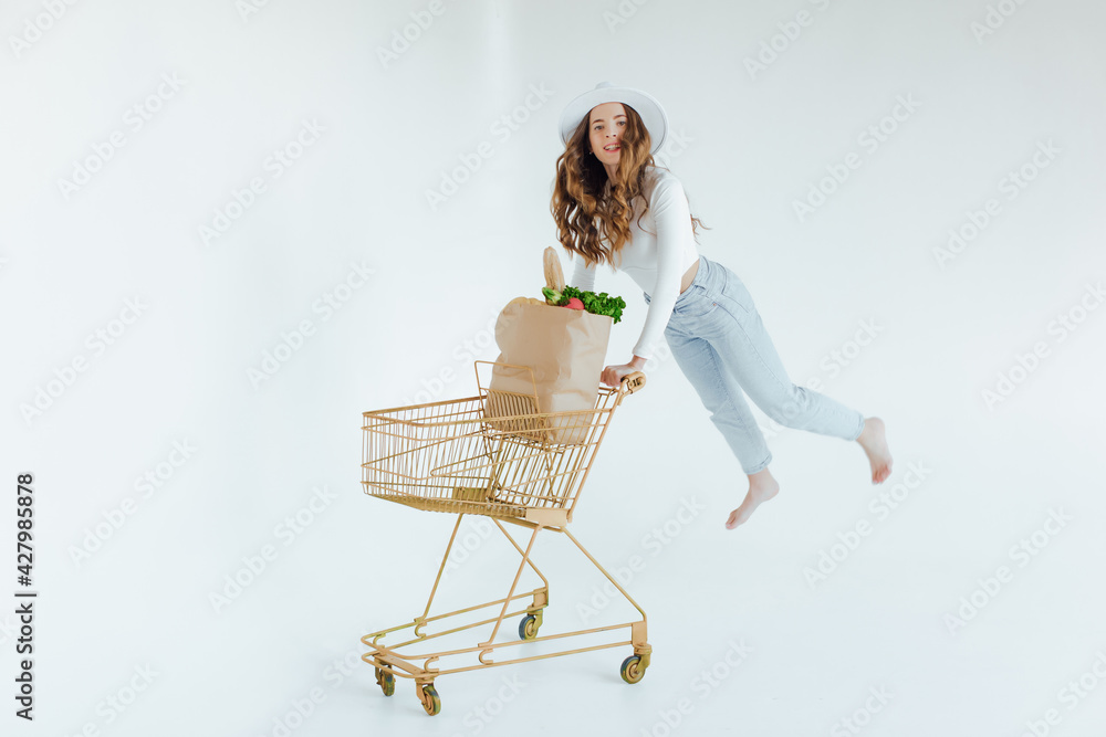 smiling young woman holding apple and looking away while standing with shopping trolley with grocery bags isolated on white