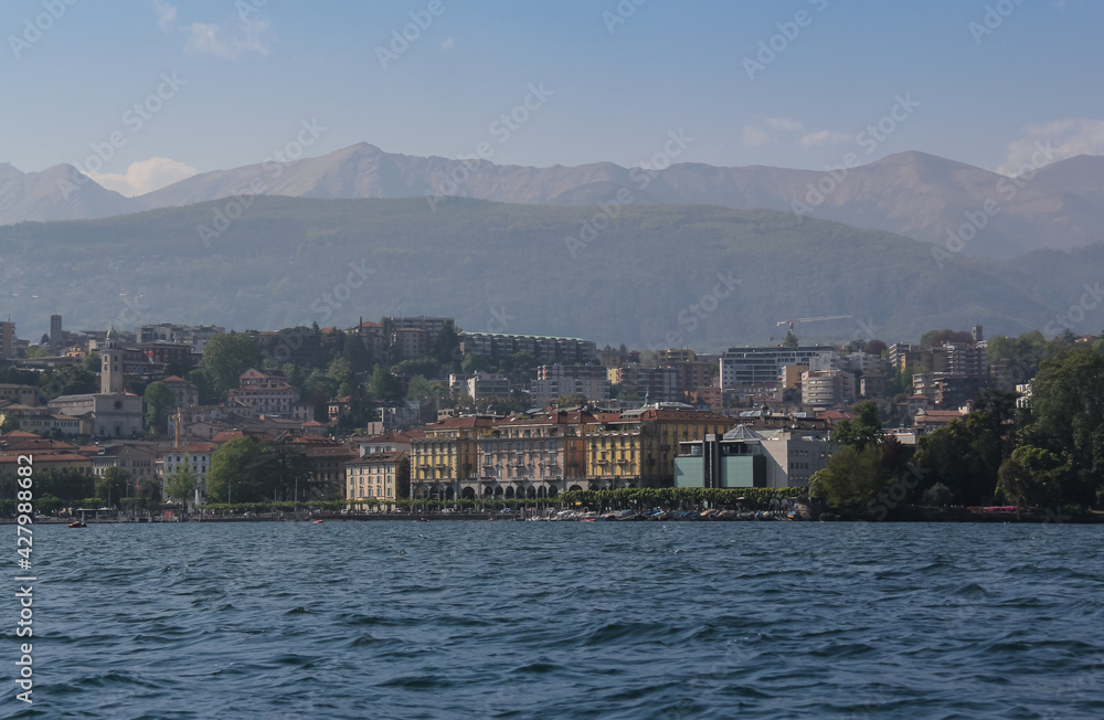 view of the city of Lugano from the lake, where you can see its buildings and the Swiss mountains in the background under a blue sky