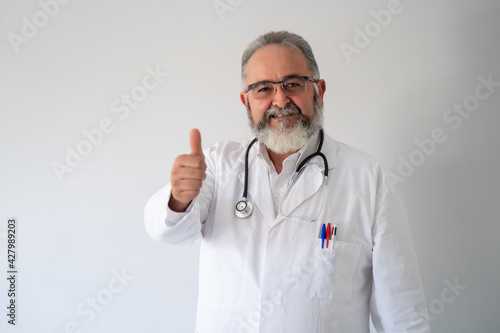 Portrait of senior bearded doctor in white coat smiling with thumbs up on white background.