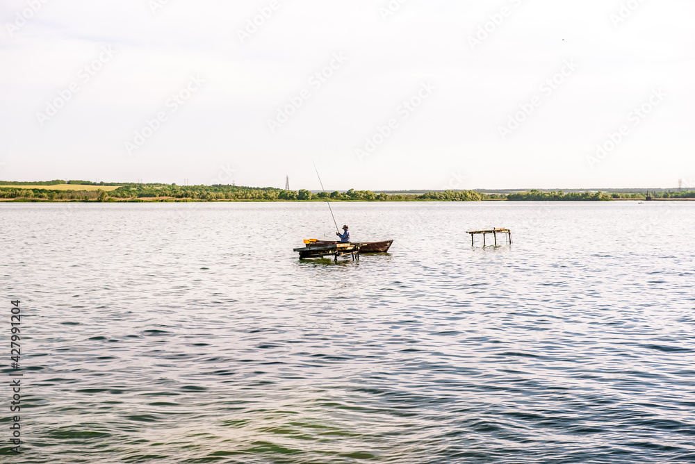 A fisherman with a fishing rod in a boat in the middle of the lake during the day. Lifestyle of a fisherman on a wooden boat to catch freshwater fish in a body of water in the middle of the day. 