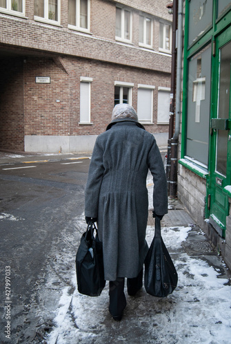 Old lady with gray coat and a plastic bag on her head. She lady walks on the snowy sidewalk holding two black bags.