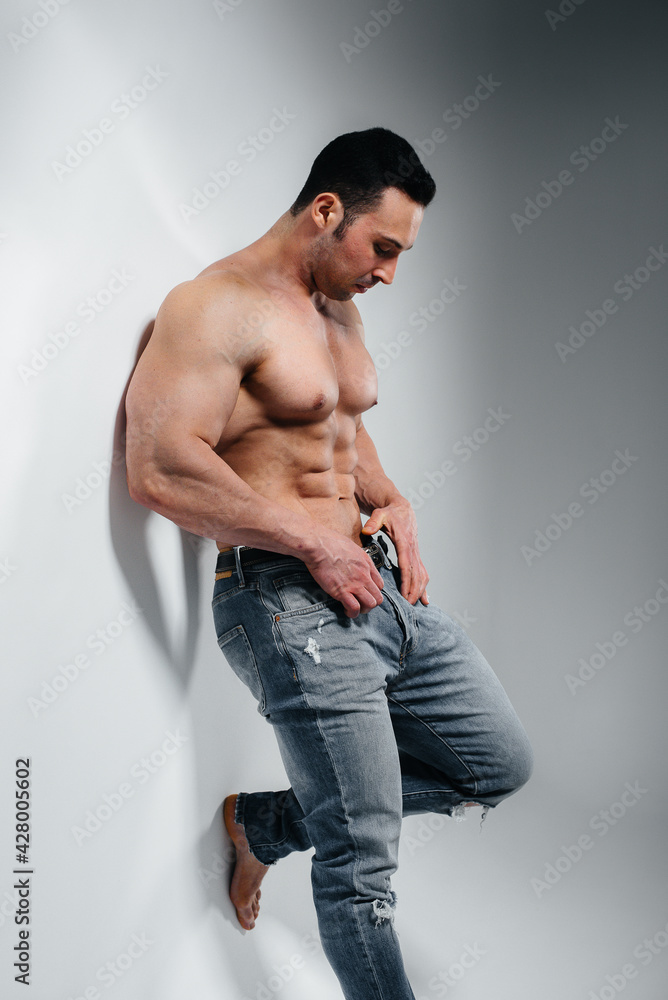 A young athlete bodybuilder poses in the studio topless in jeans near the wall. Sport. A young athlete bodybuilder poses in the studio topless in jeans near the wall. Sport.