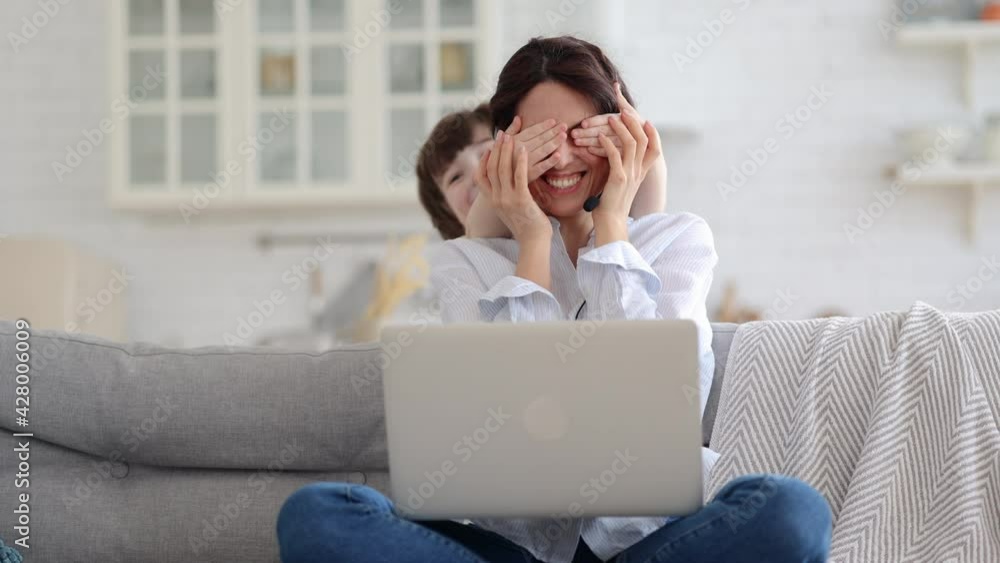 Freelancer woman sitting on couch at home office during lockdown, working on laptop. Playful child distracts from work, covering her mother's eyes, kid making noise and asking attention from busy mom. Freelancer woman sitting on couch at home office during lockdown, working on laptop. Playful child distracts from work, covering her mother's eyes, kid making noise and asking attention from busy mom.
