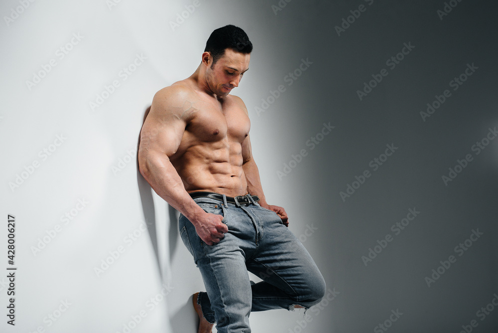 A young athlete bodybuilder poses in the studio topless in jeans near the wall. Sport. A young athlete bodybuilder poses in the studio topless in jeans near the wall. Sport.