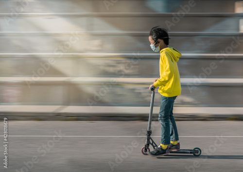 asian boy with a scooter in the park. he is wearing a mask and yellow sweater. Panning effect and selective blur.