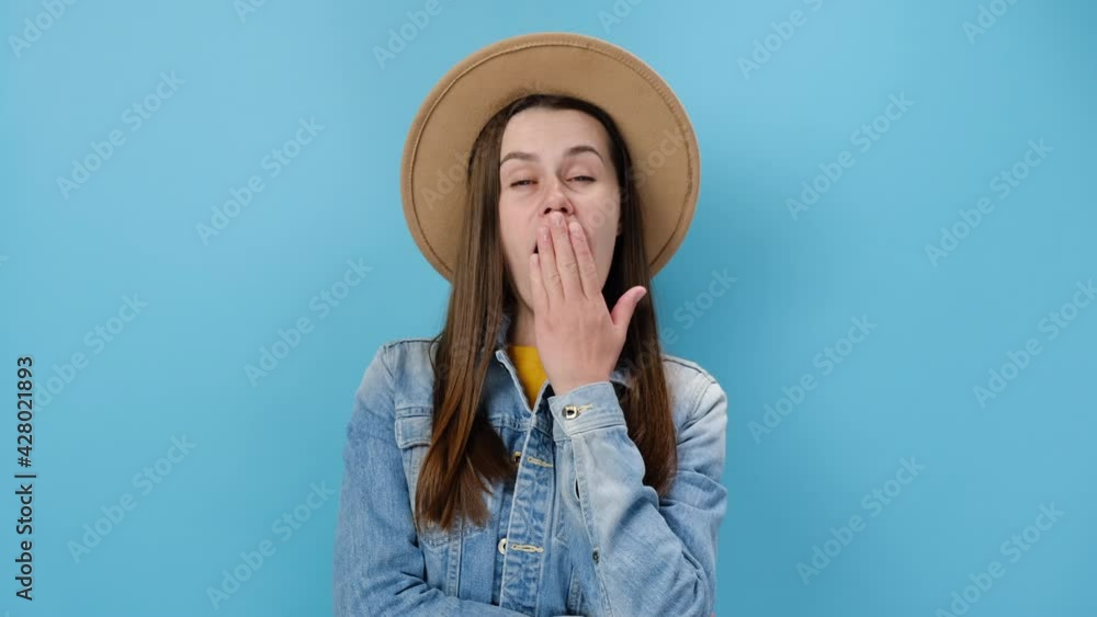 Tired young millennial female did not get enough sleep last night after party and barely got up in morning yawning, wears denim jacket and hat, isolated on blue background. People lifestyle concept Tired young millennial female did not get enough sleep last night after party and barely got up in morning yawning, wears denim jacket and hat, isolated on blue background. People lifestyle concept