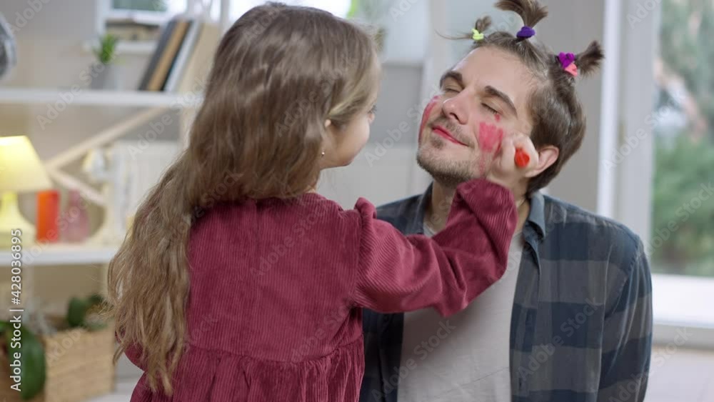 Funny man with ponytails smiling as girl painting his cheeks with red marker. Portrait of relaxed joyful Caucasian single father having fun with daughter indoors on weekend at home Funny man with ponytails smiling as girl painting his cheeks with red marker. Portrait of relaxed joyful Caucasian single father having fun with daughter indoors on weekend at home