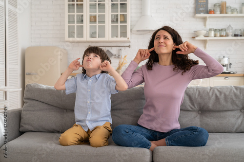 Frowning mom and son sit on couch with closed eyes and cover ears from noisy music or fight sounds from neighbors. Young female parent and kid tired of drilling noise not listen plug ears with fingers photo